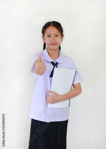 Portrait of Asian student girl in school uniform showing thumb up finger very good gesture while holding a book against white background.