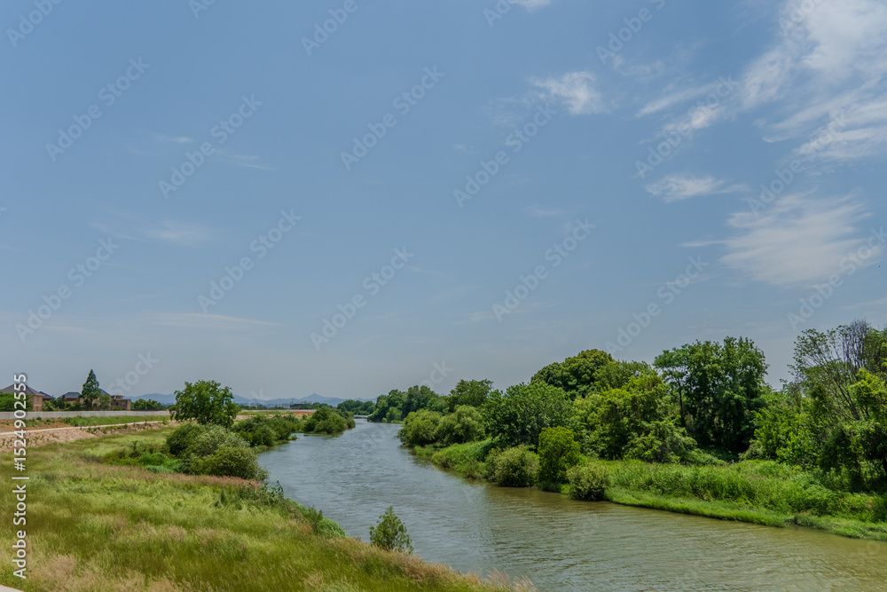 Obraz premium Winding river flowing through lush green meadows and trees under a blue sky with clouds.