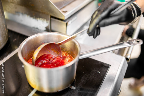Wallpaper Mural A professional chef stirs a vibrant red tomato sauce in a stainless steel pot using a ladle, wearing a black glove. The scene captures a culinary moment in a kitchen, highlighting food preparation Torontodigital.ca