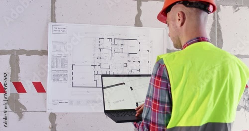 Engineer uses modern technology to study apartment layout. Builder holding computer in hands stands in front of large floor plan fixed on wall in spacious hall