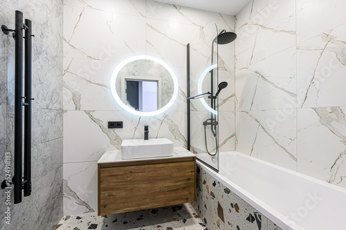 Modern bathroom featuring marble tiles, a floating wood vanity, and a round LED mirror. Black fixtures and terrazzo accents add style