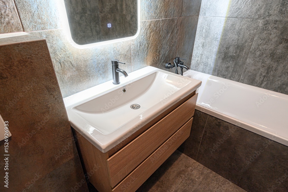 Fototapeta premium Modern bathroom with a white sink vanity, lighted mirror, and gray tile walls. Clean and contemporary design