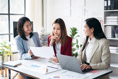 Three women are sitting at a table with papers and laptops