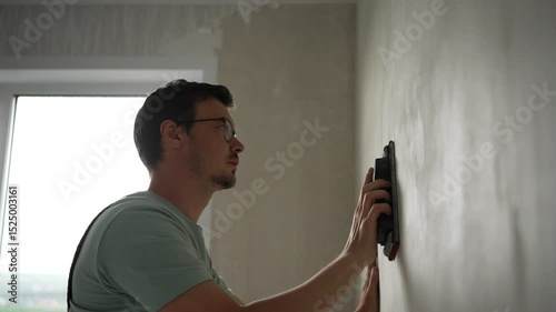 Skilled handyman leveling plastered wall surface, carefully sanding with hand tool while wearing protective work overalls and safety glasses for precise home renovation preparation