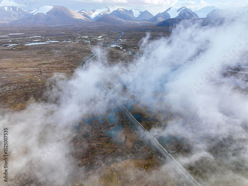An aerial view shows a winding road partially obscured by mist, stretching across a wide, rugged valley with distant mountains and water bodies