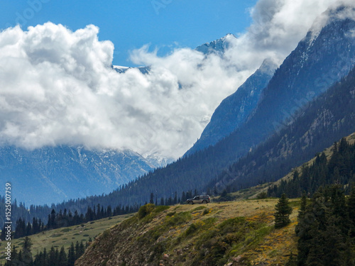 A single off-road vehicle sits on a grassy mountain ridge, overlooking a vast landscape of forests and majestic snow-capped peaks under a blue sky with clouds