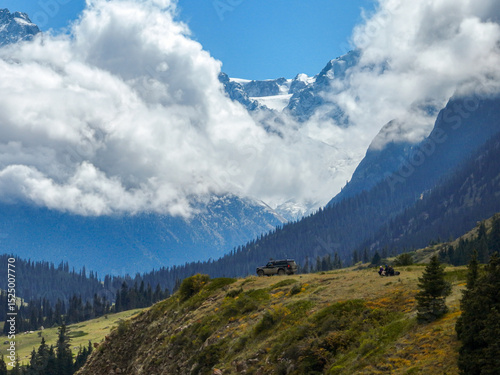 An off-road vehicle and a group of people stand on a grassy slope, surrounded by a stunning mountain landscape with snow-capped peaks and dramatic clouds