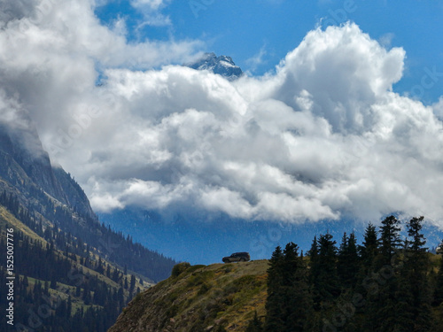 An off-road vehicle is visible on a grassy mountain slope, with dramatic clouds partially obscuring snow-capped peaks in the background