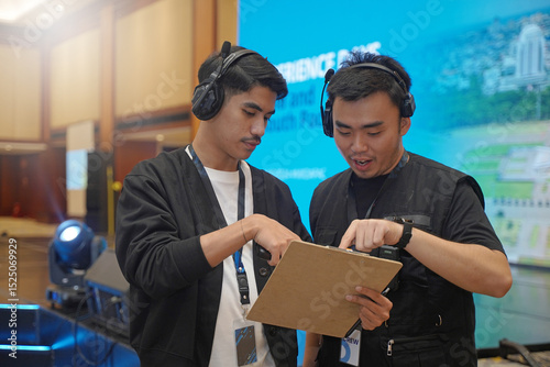 Asian male stage manager is discussing while pointing at clipboard, using headset and ID card, showing the coordination of work behind the scenes of the event with the stage atmosphere and LED screen.