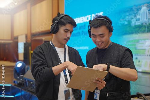 Event manager discussing rundown with stage manager while pointing at clipboard, showing behind the scenes coordination of indoor event with stage backdrop and large LED screen