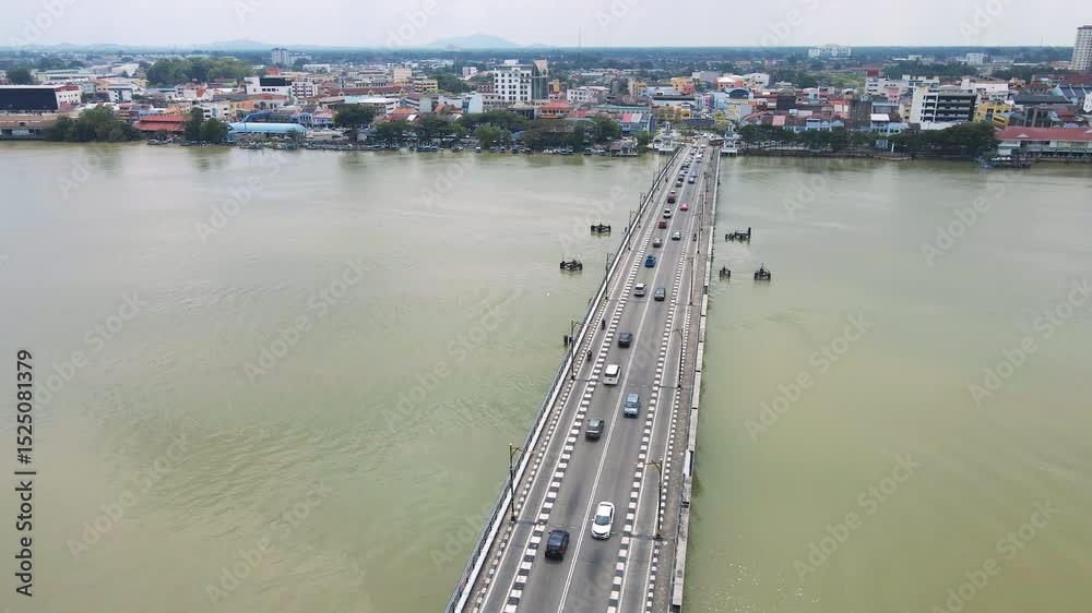 Drone view of a long bridge crossing a river with cars moving in both directions, leading into a vibrant urban town with buildings and waterfront activity. Drone view of a long bridge crossing a river with cars moving in both directions, leading into a vibrant urban town with buildings and waterfront activity.