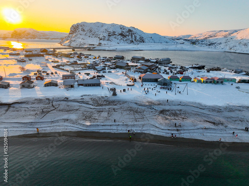 Aerial view of Teriberka village beach covered in snow, with people and colorful houses under a sunset glow, Russia