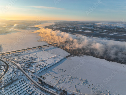 Aerial view of a wide frozen river with mist rising from it, a bridge, and snow-covered banks, in the Murmansk region, Russia