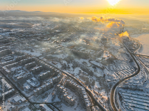 Aerial view of Murmansk city at sunset, showing residential buildings, a road, and industrial facilities with smoke plumes, Russia