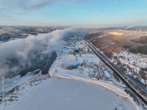 Aerial view of a winter landscape with fog rolling over a frozen river, alongside a snow-covered town and road, Russia