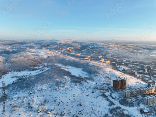 Aerial view of Murmansk, with residential buildings nestled among snow-covered hills and forests under a clear sky, Russia