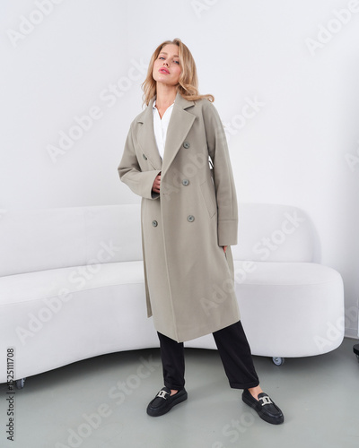 Woman wearing a stylish beige trench coat over a white shirt stands in a relaxed pose. Her hands are in the coat pockets. She's wearing black pants and loafers, in a modern interior