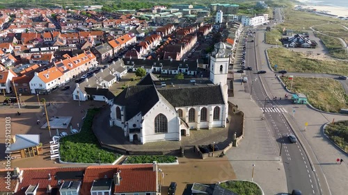 Aerial view of a Dutch town with a prominent church in the center, surrounded by residential homes, green trees, and a clear blue sky on a bright spring day. Peaceful and charming atmosphere.