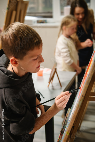 A school boy drawing brush landscapes on easel. Teacher explains to kid how to draw pictures in art class. Child enjoys painting in workshop. Art lesson. Education, creativity, learning concept.