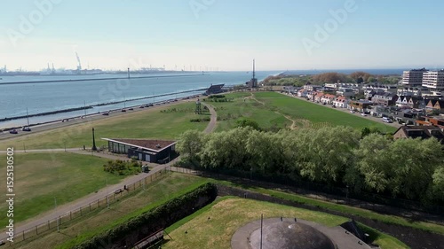 A coastal view showing a quiet seaside town with a rocky shoreline, green open fields, a road with passing vehicles, and residential buildings in the distance under a clear blue sky. Windmills.
