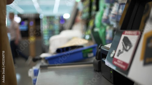 Customer hand taking a paper receipt from a self-service checkout machine in a brightly lit hardware store, with blurred customers and shelves in the background
