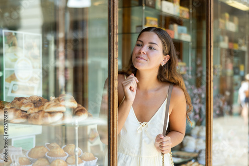 Girl in summer clothes stopped near display case with sweets, examines view and chooses sweet snack, makes order for cookies, cupcakes in bakery