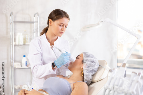 Fotografie Professional female aesthetician administering hardware procedure on young woman in cosmetology clinic, using ice hammer attachment to relieve inflammation and improve tone of client facial skin