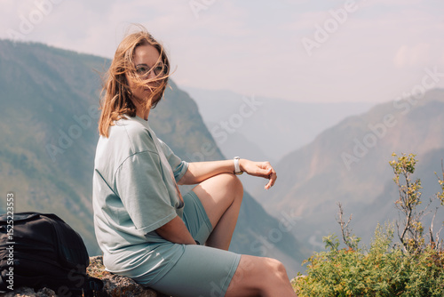 Young woman in sportswear sits on a rock ledge, peacefully observing scenic mountain landscape during a summer hiking adventure.