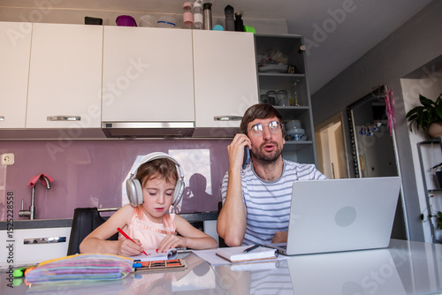 Busy father working remotely from home using laptop and having phone call while his little daughter drawing and listening to music on headphones in kitchen