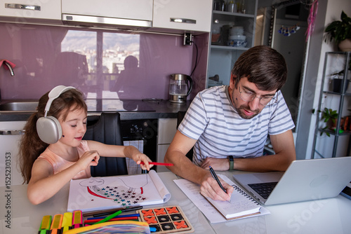 Father working from home on a laptop while daughter sits beside him, drawing a rainbow with colored pencils and enjoying music through headphones at the kitchen table