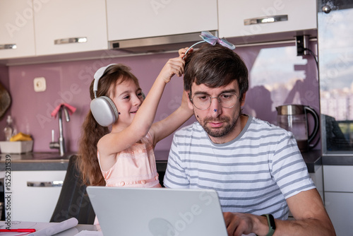 Daughter wearing headphones is playing with her father trying to put a toy on his head while he's working from home using a laptop in the kitchen