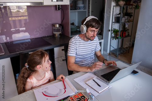 Father wearing headphones and working on a laptop while his young daughter draws at the kitchen table in a modern apartment, creating a warm atmosphere of family togetherness