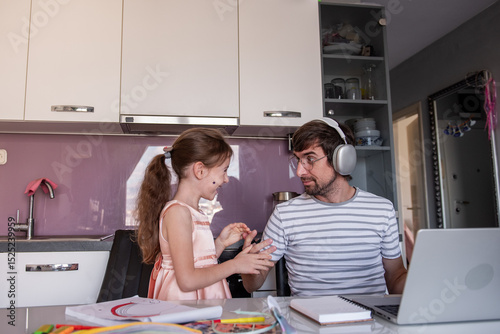 Young father wearing headphones, working on his laptop in the kitchen while his little daughter interrupts with playful chatter and laughter, creating a lively family atmosphere