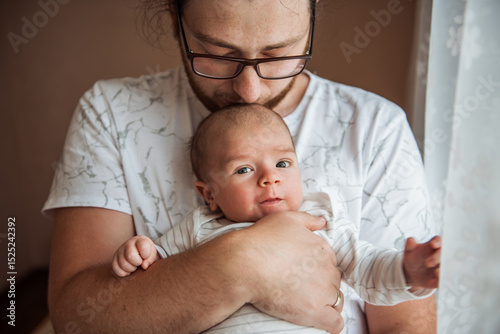 Tender moment as a father cradles his newborn baby near a window. The soft lighting highlights the bond and warmth shared between them, capturing the essence of parenthood