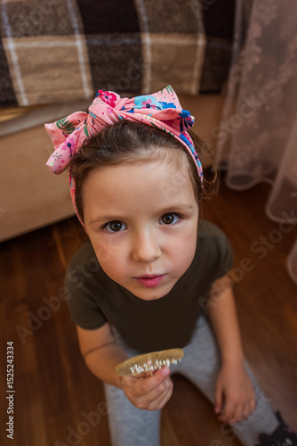 Little girl wearing a pink headband, sitting on the floor of her cozy bedroom, holding a wooden comb and looking up with curiosity and joy, embodying childhood innocence and playfulness