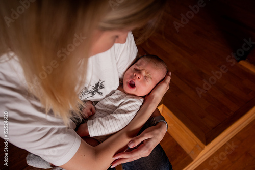 Young mother cradling her yawning newborn baby in her arms during a quiet moment at home. Natural indoor lighting creates an intimate atmosphere while the infant displays signs of tiredness