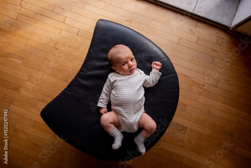 Newborn baby lying comfortably on a crescent-shaped black cushion. The wooden floor adds warmth to the scene, highlighting the baby's peaceful expression and cozy outfit