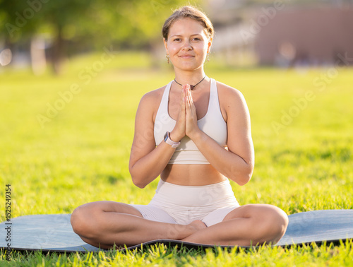 Smiling young girl meditating in lotus position with hands clasped in namaste during serene morning yoga practice on green sunlit meadow, harmonizing breath and calming against summer landscape
