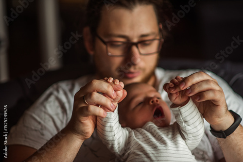 Father lovingly holds his newborn baby's hands, gently lifting them while the baby yawns. The intimate moment captures the warmth and connection of early parenthood in a cozy home setting