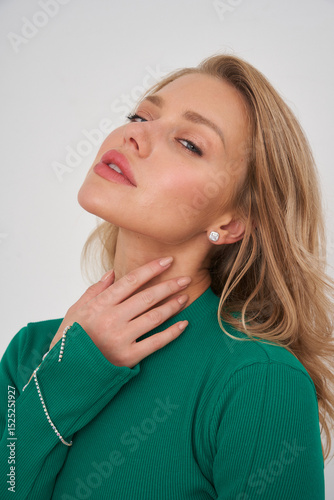 Elegant woman with long blonde hair poses in a green ribbed top, hand touching her neck, wearing sparkly earrings. Her expression is confident, with subtle eyeshadow and pink lips