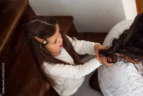 Young girl with a hair clip carefully braiding the hair of a seated man on wooden stairs. The scene captures a moment of bonding and creativity in a cozy home environment