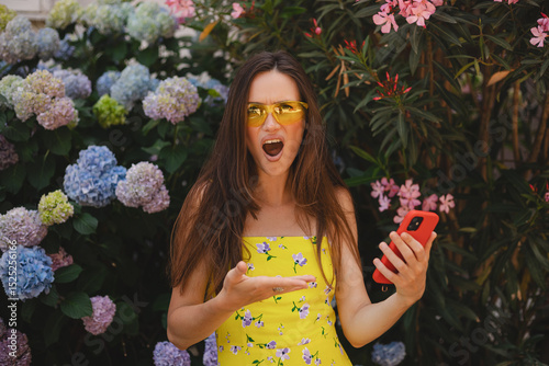 Mad and shock young brunette beautiful woman using mobile phone standing near blossom hydrangea. Unhappy woman hold mobile phone. Disappointed sad upset lady horrified impressed news. WTF. Oh no.