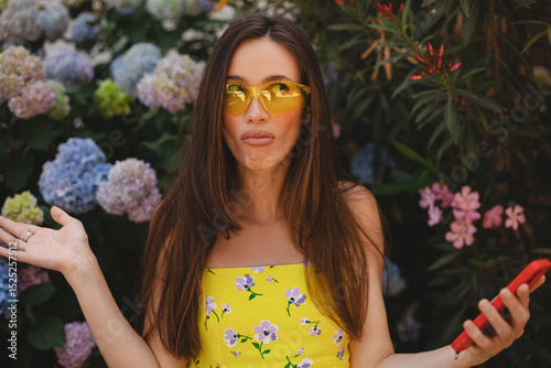 Mad and shock young brunette beautiful woman using mobile phone standing near blossom hydrangea. Unhappy woman hold mobile phone. Disappointed sad upset lady horrified impressed news. WTF. Oh no.