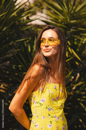 Brunette long hair woman standing near palm tree and look coquette back, flirty smiling. Woman wear yellow dress in flower print and yellow sunglasses, summer mood.