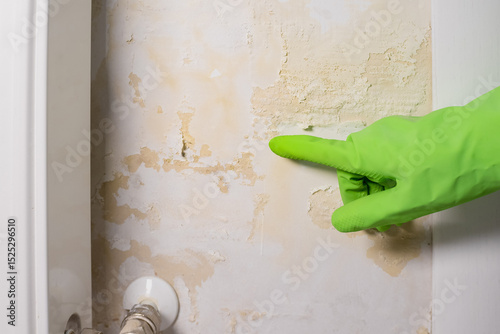 Woman in gloves showing flooded wall. The wall is peeling near the heating radiator. Pipe leak problem. Renovation concept.