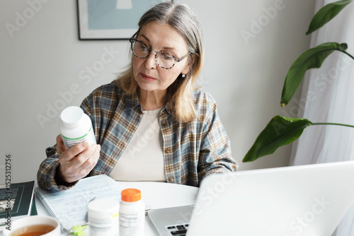 Aged woman holding bottle of dietary supplement and reading label with instruction, ingredients or brand name carefully before ordering vitamins online using laptop and web application
