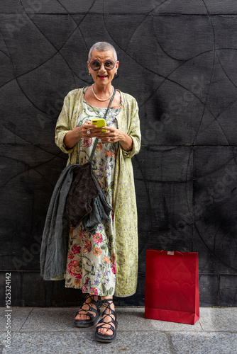 elegant smiling mature woman with shaved grey hair texting on cell phone in front of black wall