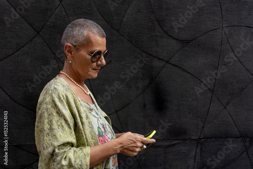 elegant smiling mature woman with shaved grey hair texting on cell phone in front of black wall