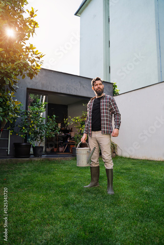 Gardener standing proudly on freshly cut lawn with watering can in his hand, sun coming through tree branches in background 
