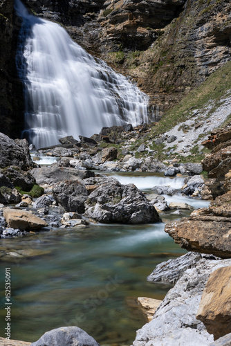 Long exposure of Cola de Caballo waterfall flowing into a clear turquoise river surrounded by rocks and vegetation in Ordesa y Monte Perdido National Park, Spain